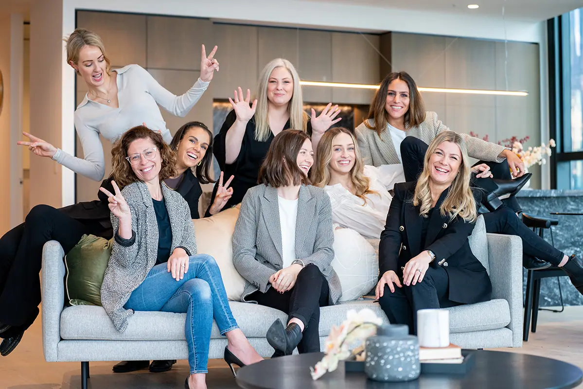 A team of eight women posing playfully together on and around a lounge in a modern office.