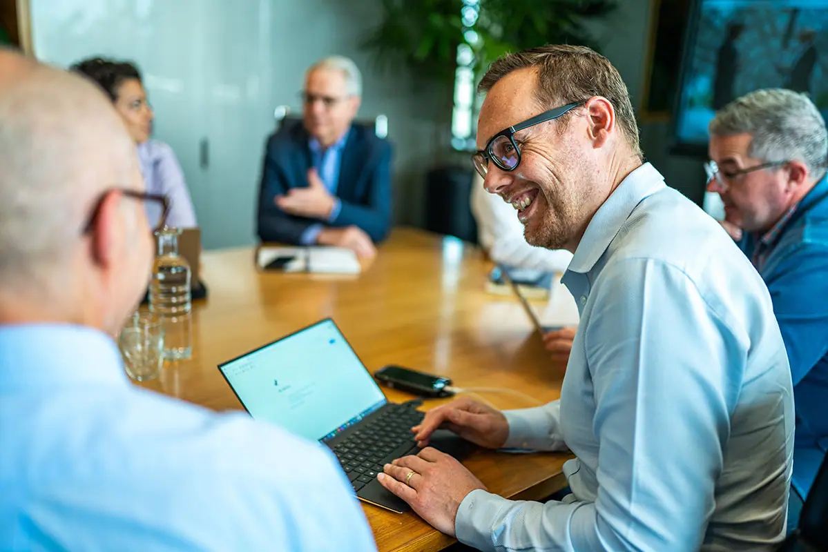 A man laughing during a boardroom meeting while working on a laptop, colleagues seated around the table.