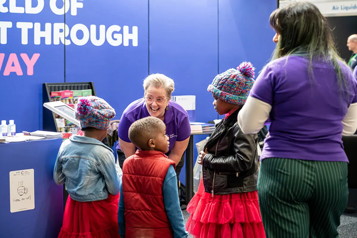 A staff member in a purple uniform crouching to talk with young children at a community health expo stand.