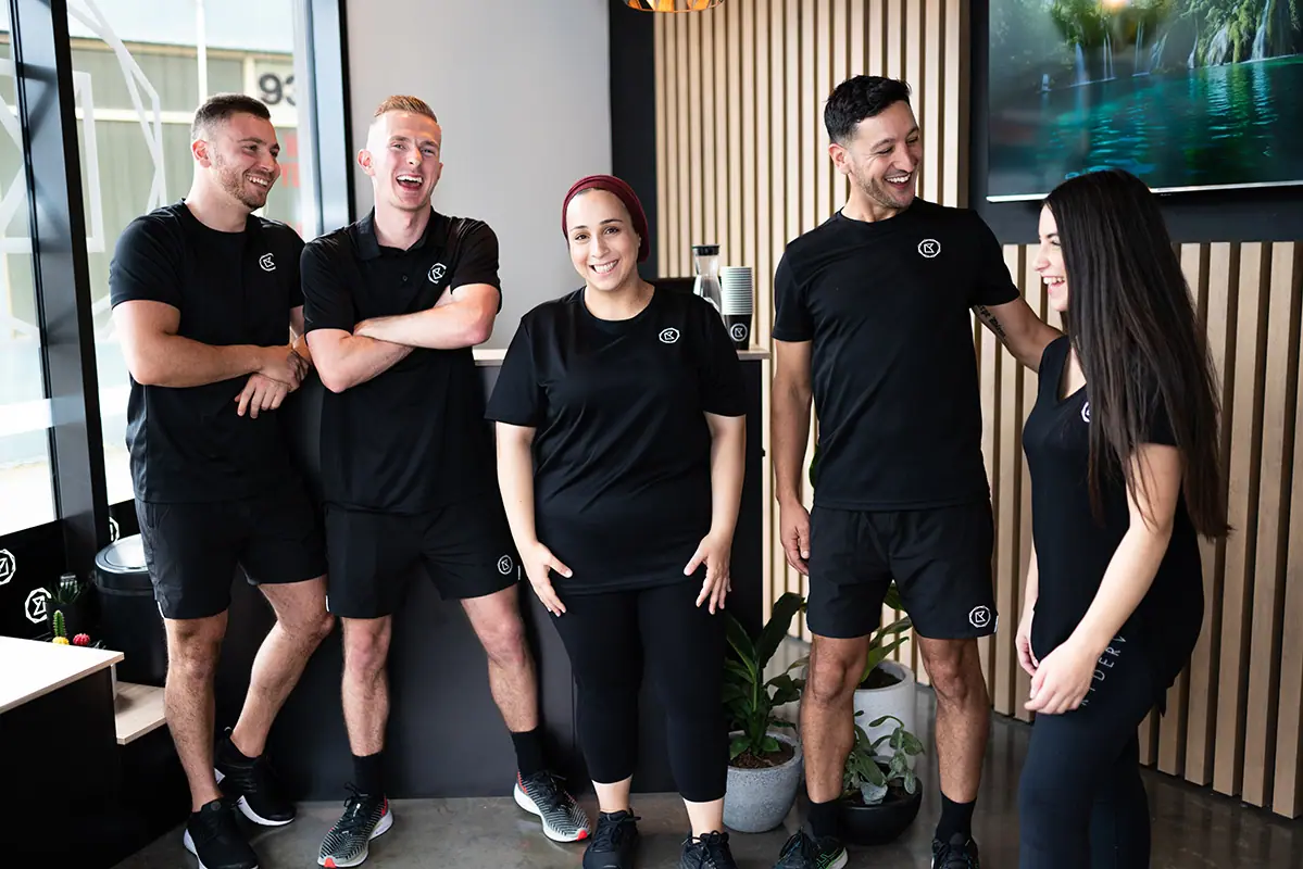 Five gym staff in matching black uniforms laughing together in a fitness studio reception.