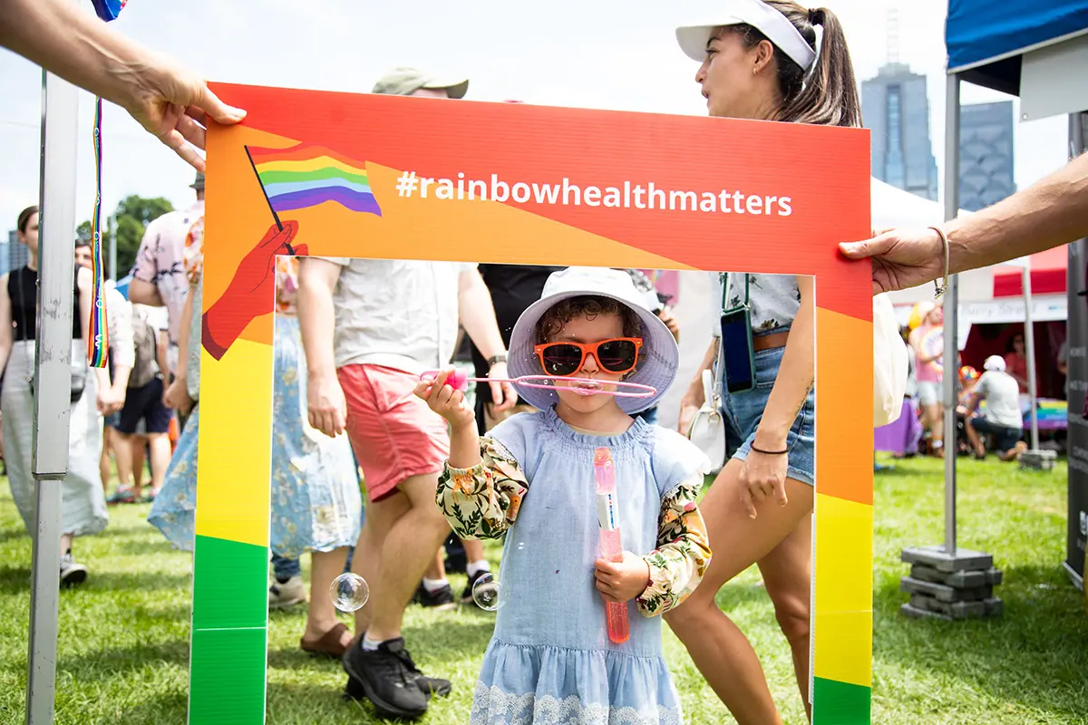A young child blowing bubbles while standing inside a colourful #rainbowhealthmatters photo frame at an outdoor festival.