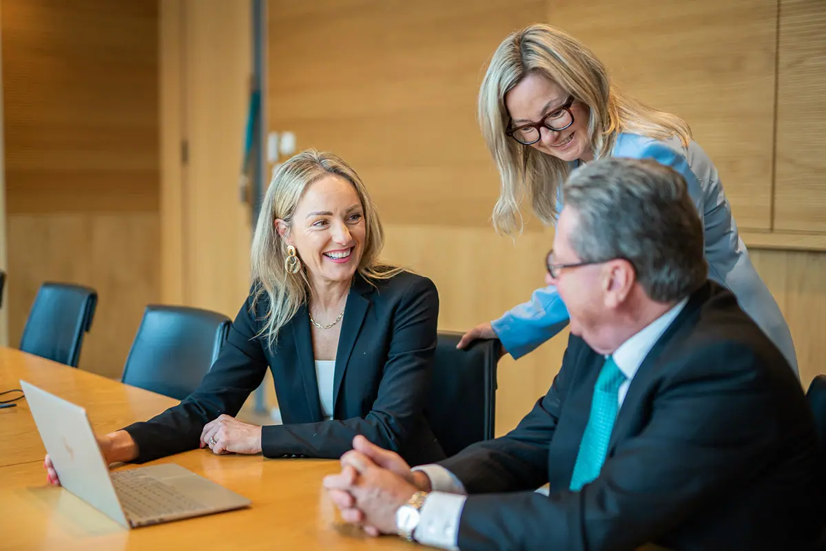Three colleagues in business dress laughing together over a laptop in a timber-panelled boardroom.